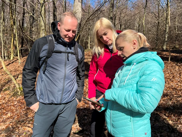 Three people in an autumnal forest, focused on a shared cell phone; blue sky in the background.