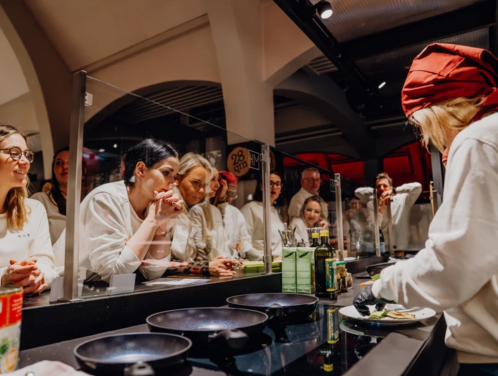 MarkthalleHerford-66.jpg Eine Gruppe von Personen beobachtet einen Koch in roter Mütze, der hinter Glas an einem Herd arbeitet.