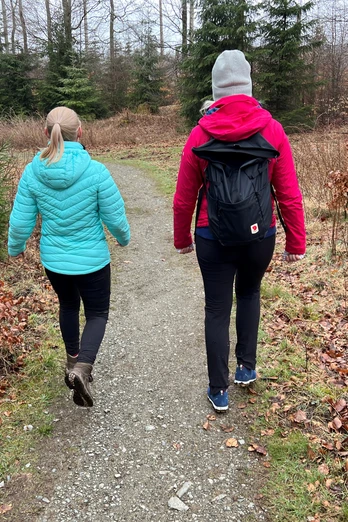 Zwei Personen spazieren auf einem schmalen Waldweg, umgeben von kahlen Bäumen mit leichtem Laub.