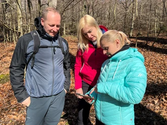 Three people in the forest looking at a map in sunny weather; all wearing outdoor clothing.