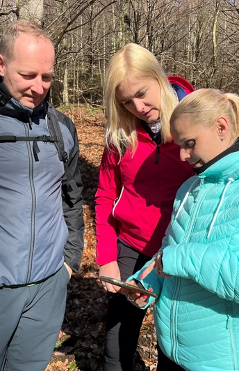 Drei Personen im Wald betrachten eine Karte bei sonnigem Wetter; alle tragen Outdoor-Kleidung.