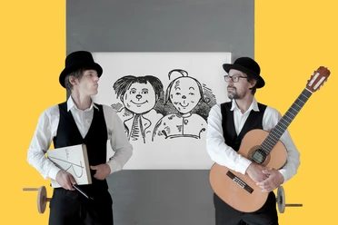 Zwei Männer in schwarzen Westen und Hüten stehen lächelnd vor einer Zeichnung, einer spielt Gitarre.