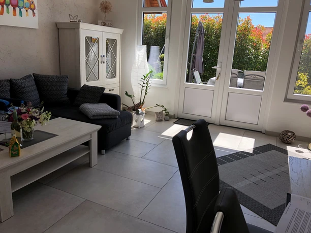 Zwetschenbrummer Helles Wohnzimmer mit Glastüren, modern eingerichtet. Ein dunkles Sofa und Pflanzen verleihen Flair.