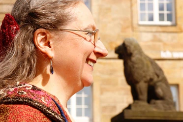 Löwenpudelmittel.jpg Portrait einer lächelnden Frau im Profil mit einem steinernen Löwen im Hintergrund bei Sonnenschein.