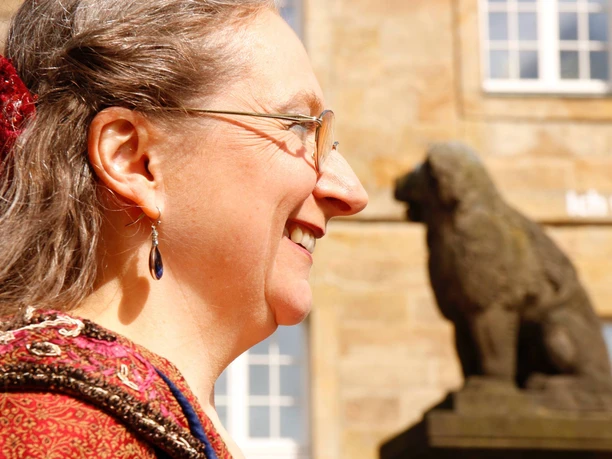 Portrait einer lächelnden Frau im Profil mit einem steinernen Löwen im Hintergrund bei Sonnenschein.