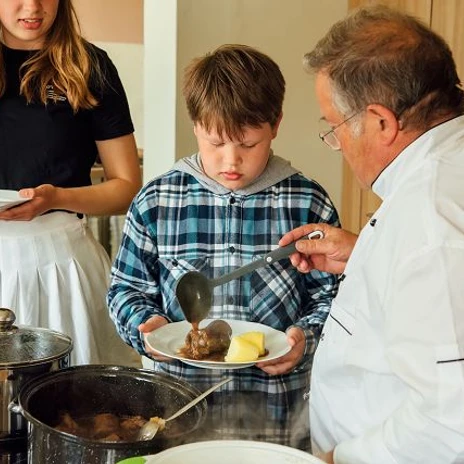 k-blankenhain-rouladen-machen-10-c-tmgs-czech-vibes.jpg Ein Junge erhält von einem Koch Rouladen mit Kartoffeln auf einen Teller, während eine Frau zusieht.