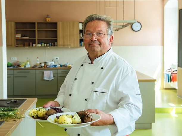 k-blankenhain-rouladen-machen-13-c-tmgs-czech-vibess.jpg Ein Koch in weißer Uniform präsentiert Rouladen und Kartoffeln auf Tellern in einer Küche.