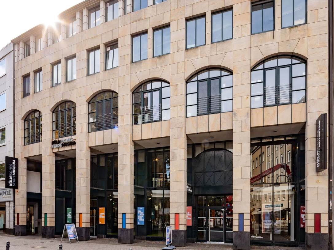 Cologne City Museum Das Bild zeigt die Fassade des Kölnischen Stadtmuseums, ein modernes Gebäude mit großen Fenstern und hellen Steinwänden. Der Eingang ist zur Straße hin ausgerichtet, flankiert von Informationsschildern und Plakaten, die auf aktuelle Ausstellungen hinweisen. The picture shows the façade of the Cologne City Museum, a modern building with large windows and light-colored stone walls. The entrance faces the street, flanked by information signs and posters advertising current exhibitions.