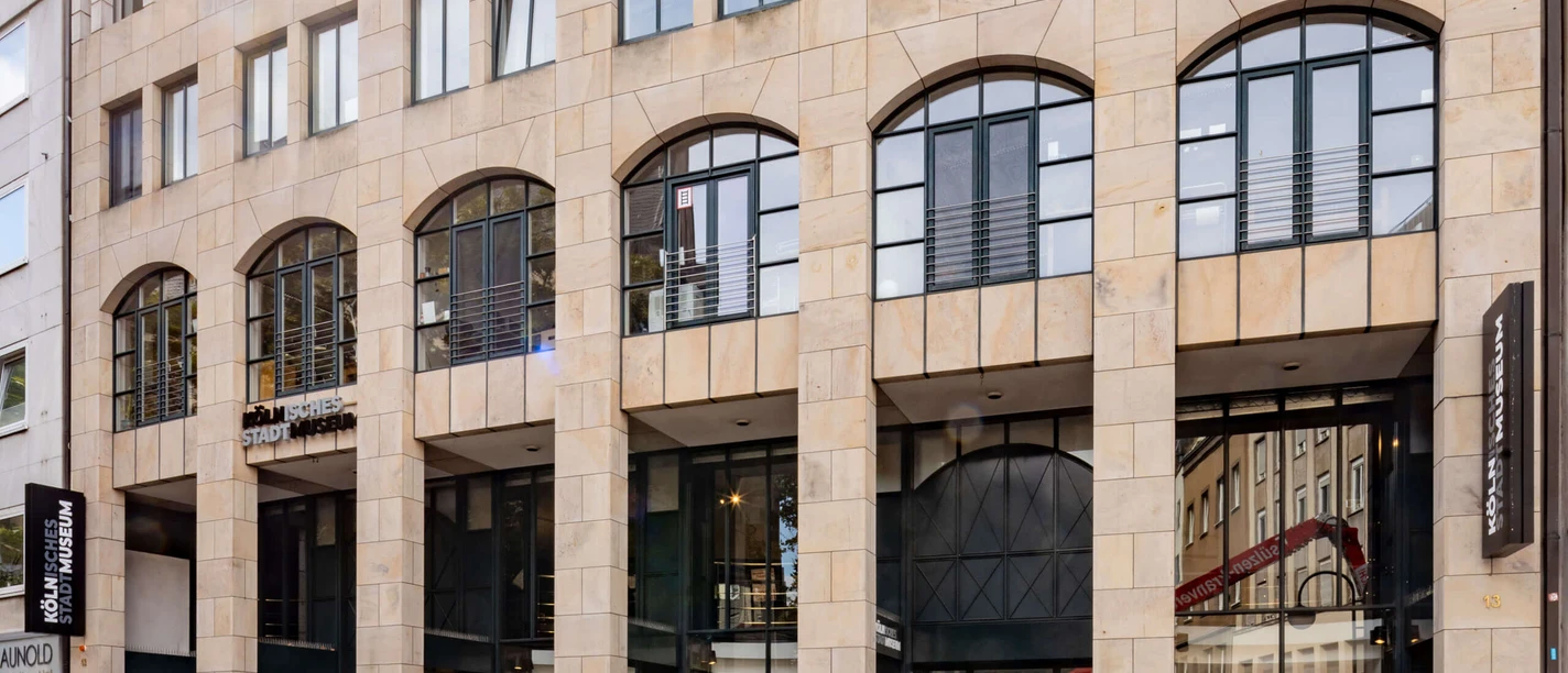 Kölnisches Stadtmuseum Das Bild zeigt die Fassade des Kölnischen Stadtmuseums, ein modernes Gebäude mit großen Fenstern und hellen Steinwänden. Der Eingang ist zur Straße hin ausgerichtet, flankiert von Informationsschildern und Plakaten, die auf aktuelle Ausstellungen hinweisen.