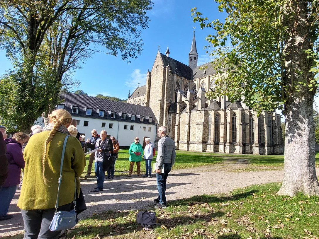 LuGeV-Führung in Altenberg Gruppe von Menschen steht vor einer historischen Kirche aus Stein an einem sonnigen Tag, umgeben von Bäumen.