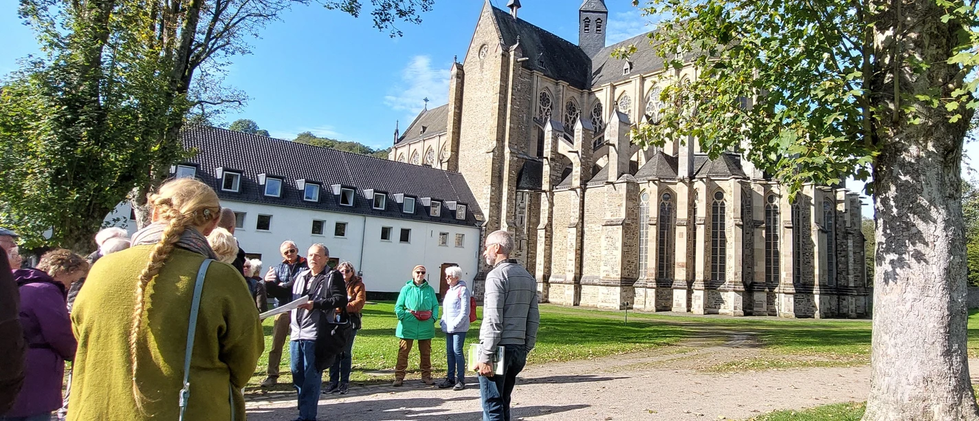 LuGeV-Führung in Altenberg Gruppe von Menschen steht vor einer historischen Kirche aus Stein an einem sonnigen Tag, umgeben von Bäumen.