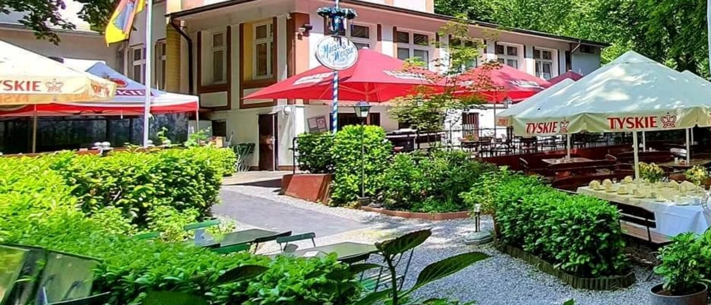 Biergarten in der Schlesischen Schänke im Engelsberger Hof Biergarten der Schlesischen Schänke in Solingen, umgeben von Bäumen, mit bunten Schirmen und Sitzflächen.