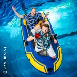 Zwei lachende Personen in Hawaii-Hemden treiben in einem Schlauchboot auf klarem, blauem Wasser.