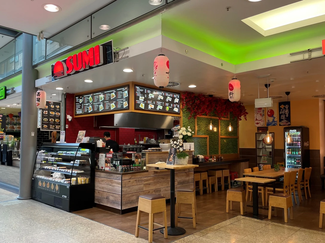 Sumi.jpg Ansicht des Restaurants bzw. des Bedientresens von vorne. Rechts daneben sieht man einen Sitzbereich mit Tischen und Stühlen.