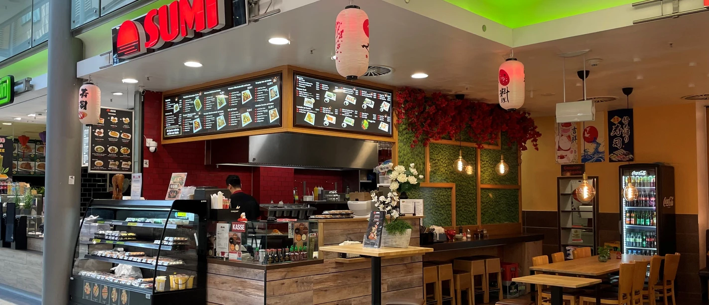 Sumi.jpg Ansicht des Restaurants bzw. des Bedientresens von vorne. Rechts daneben sieht man einen Sitzbereich mit Tischen und Stühlen.