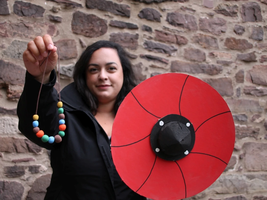 Schild_und_Kette_KLEIN_Gizem_Cevik_C_Huxarium_Gartenpark_Höxter.jpg Frau zeigt Schild und Perlenkette