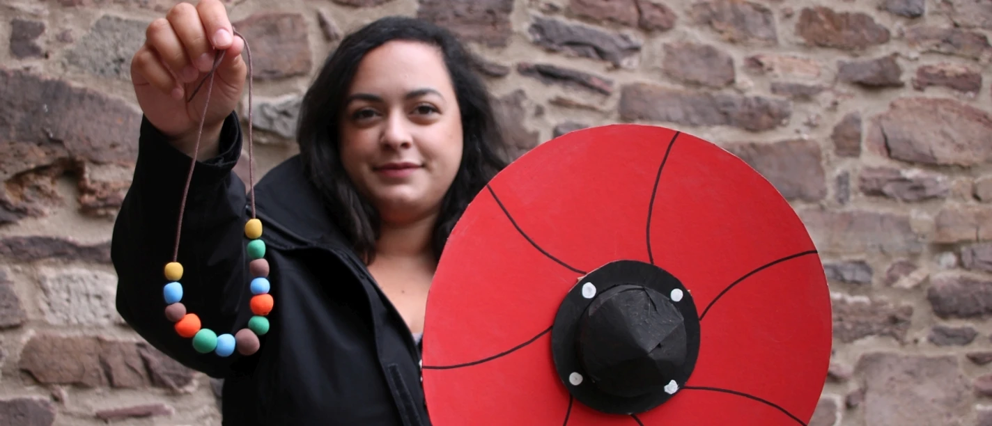 Schild_und_Kette_KLEIN_Gizem_Cevik_C_Huxarium_Gartenpark_Höxter.jpg Frau zeigt Schild und Perlenkette