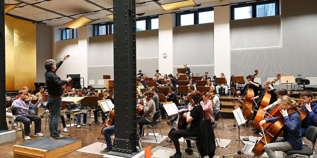 Jugendsinfonieorchester Aachen (c) Sandra Borchers.jpg Junge Musiker*innen sitzen in einem hohen Raum und musizieren, dirigiert von einem Mann in Hemd und Jeans.