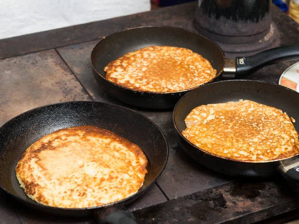 buttermilchplinsen-schleife-c-andreas-krone-cc0.jpg Drei Pfannen mit frisch gebackenen Plinsen auf einem Herd in einer rustikalen Küche.