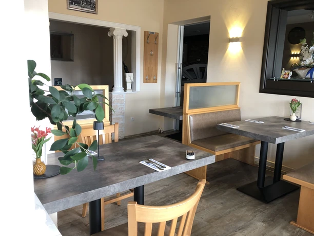 Restaurant Olympia Einladender Restaurant-Innenbereich mit Holzmöbeln, grünen Pflanzen und sanfter Wandbeleuchtung.