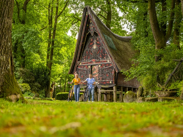 Batakhaus, Werpeloh – Radfahren Hümmling ©Emsland Tourismus GmbH (1).jpg Zwei Radfahrende vor einem reich verzierten Batakhaus im Grünen von Werpeloh im Emsland