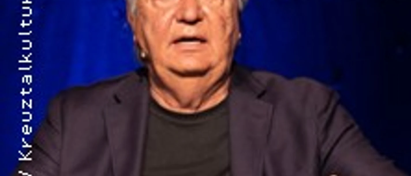 Älterer Mann auf Bühne vor blauem Hintergrund in sprechender Gestik.