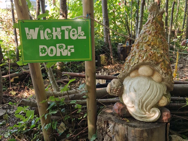 NaRa Wichteldorf Wichtel aus Holz, trägt spitze Mütze, steht neben Schild im Wald, auf dem 'Wichteldorf' steht.