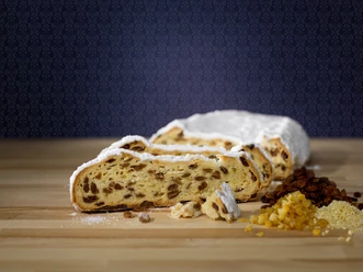 stollen-geschnitten-zutaten-c-schutzverband-dresdner-stollen-e-v-katharina-grottker.jpg Ein angeschnittener Stollen auf Holzbrett, umgeben von Rosinen und kandierten Früchten.
