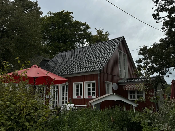 Hofcafé Kaffeekännchen Gemütliches rotes Holzhaus mit weißen Fenstern, umgeben von grünen Bäumen und einem roten Sonnenschirm.