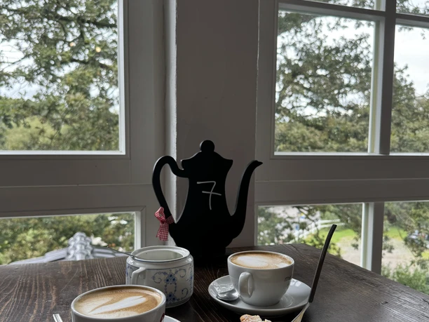 Kaffee & Käsekuchen Kaffeeszene mit Cappuccinos, Teekanne und Gebäck, vor großen Fenstern mit grünem Ausblick.