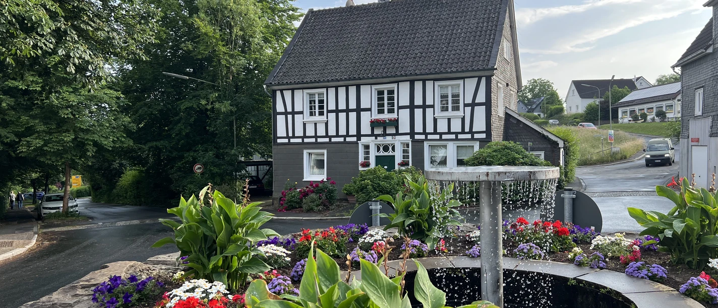 Ortskern Nümbrecht Ein traditionelles Fachwerkhaus mit blühendem Blumenbeet und zentralem Springbrunnen in einer ländlichen Umgebung.