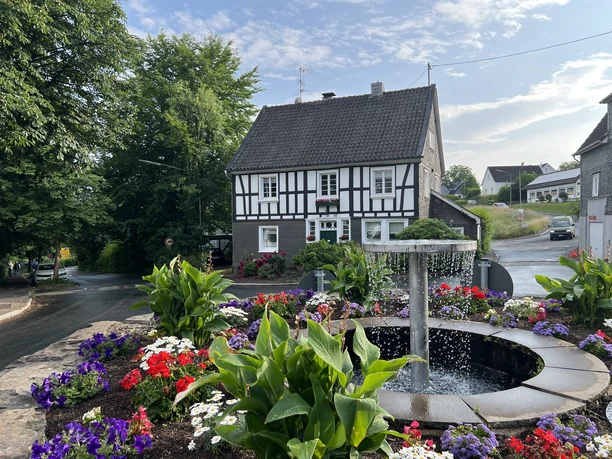 Ortskern Nümbrecht Ein traditionelles Fachwerkhaus mit blühendem Blumenbeet und zentralem Springbrunnen in einer ländlichen Umgebung.