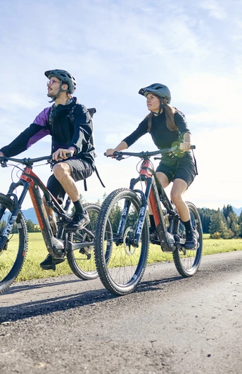 Radpilgern-Klöster und Natur entdecken Zwei Radfahrer auf einem Weg entlang grüner Felder, mit Bergen im Hintergrund.