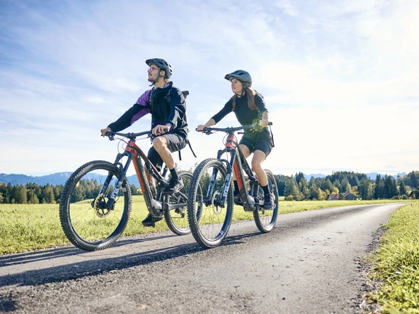 Radpilgern-Klöster und Natur entdecken Zwei Radfahrer auf einem Weg entlang grüner Felder, mit Bergen im Hintergrund.
