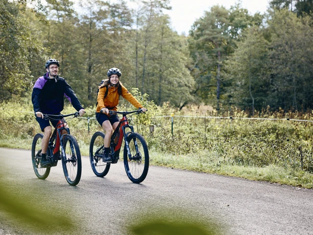 Radpilgern-Kraftquelle Zwei Menschen fahren mit Mountainbikes auf einem Weg, umgeben von Bäumen.