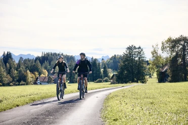 Radpilgern - Stille Wege Zwei Radfahrer auf einem asphaltierten Weg durch ländliche Landschaft mit Wiesen und Bäumen.