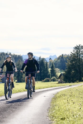 Radpilgern - Stille Wege Zwei Radfahrer auf einem asphaltierten Weg durch ländliche Landschaft mit Wiesen und Bäumen.