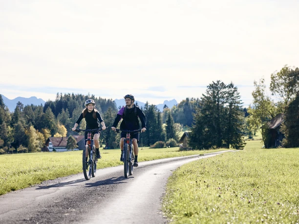Radpilgern - Stille Wege Zwei Radfahrer auf einem asphaltierten Weg durch ländliche Landschaft mit Wiesen und Bäumen.