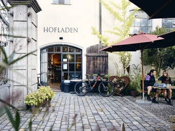 Radpilgern - Hofladen Bernried Hofladen mit geöffneter Tür, Fahrrad davor, zwei Personen sitzen unter Sonnenschirm.