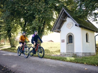 Radpilgern - Impulstafel Polling Zwei Personen radeln auf einem Weg neben einer kleinen Kapelle unter Bäumen.