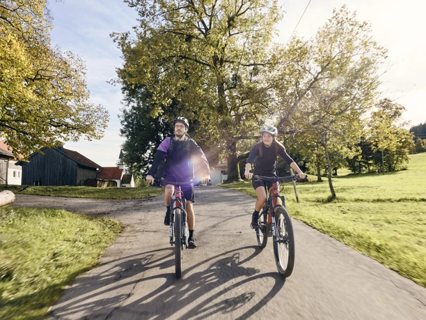 Radpilgern - Auerberg Bernbeuren Zwei Radfahrer auf einem Landweg bei sonnigem Wetter, umgeben von grüner Landschaft.