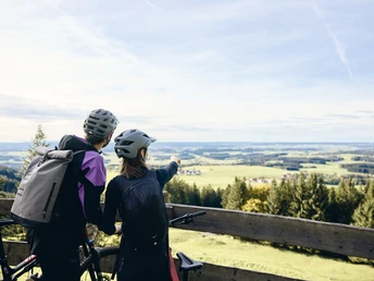 Radpilgern - Auf dem Auerberg Zwei Radfahrer mit Helmen genießen den Blick von einem Holzgeländer auf eine weite Landschaft.