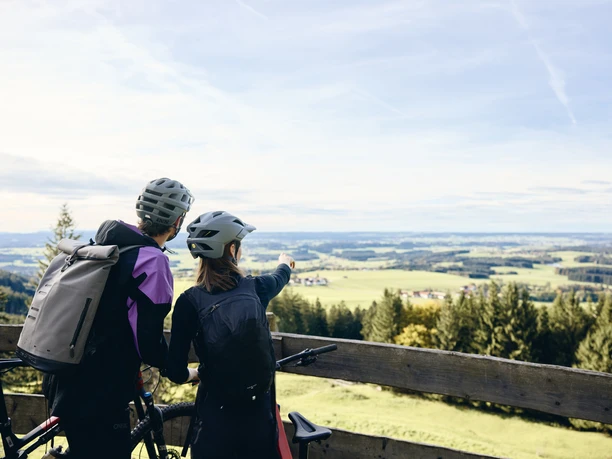 Radpilgern - Auf dem Auerberg Zwei Radfahrer mit Helmen genießen den Blick von einem Holzgeländer auf eine weite Landschaft.