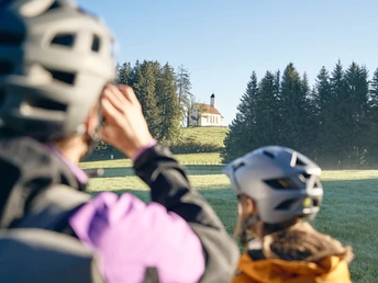 Radpilgern - Heilig-Kreuz-Kirche Steingaden Menschen mit Fahrradhelmen betrachten die Heilig-Kreuz-Kirche auf einem Hügel.