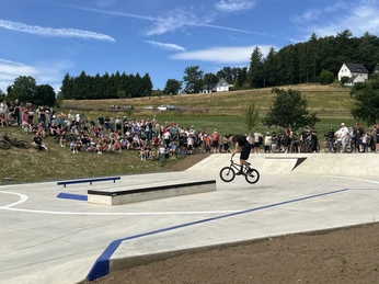 Skate und Bikepark Waldbröl Eine große Menschengruppe beobachtet BMX-Fahrer auf einer Beton-Skateanlage vor grüner Landschaft.
