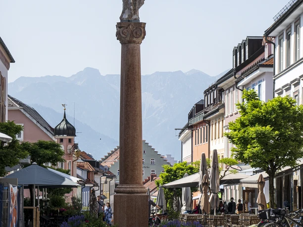 Fußgängerzone Murnau Fußgängerzone in Murnau, Statue auf Säule, Berge im Hintergrund, Straßencafés und Radfahrer.
