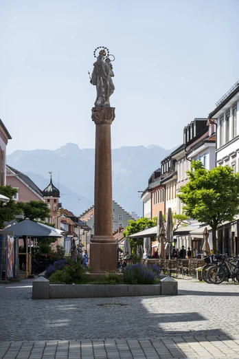 Himmlisch genießen - Murnau Fußgängerzone in Murnau, Statue auf Säule, Berge im Hintergrund, Straßencafés und Radfahrer.
