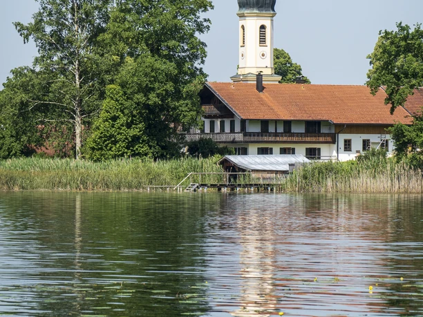 Himmlisch genießen - See Kirchturm und Gebäude spiegeln sich im ruhigen See, umgeben von grünen Bäumen.