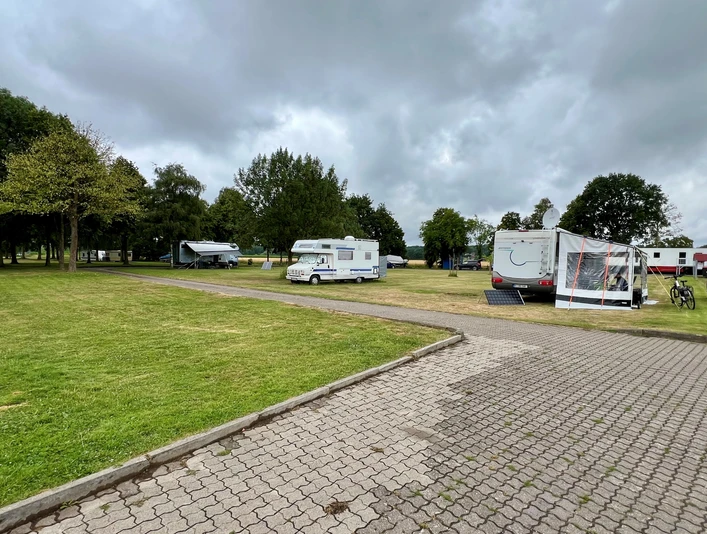 Wohnmobilstellplatz Heisterholz Grüne Wiese mit mehreren geparkten Wohnmobilen unter bewölktem Himmel, umgeben von Bäumen.