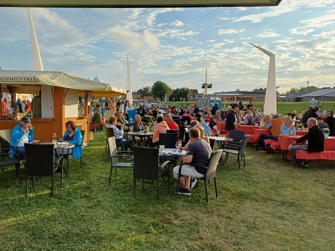 Weinfest Greetsiel Menschen sitzen an Tischen auf der Wiese am Hafen von Greetsiel und genießen Wein bei Abendsonne.
